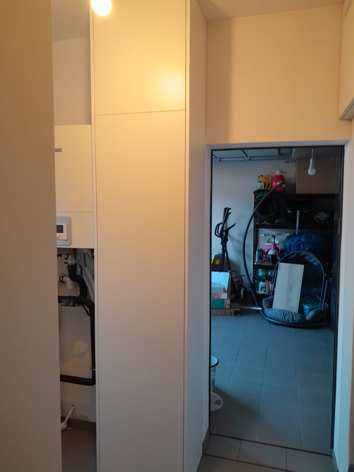 Biała szafa w przedpokoju, z widoczną instalacją hydrauliczną za otwartymi drzwiami i widokiem na zagracony garaż przez otwarte drzwi po prawej stronie szafy.