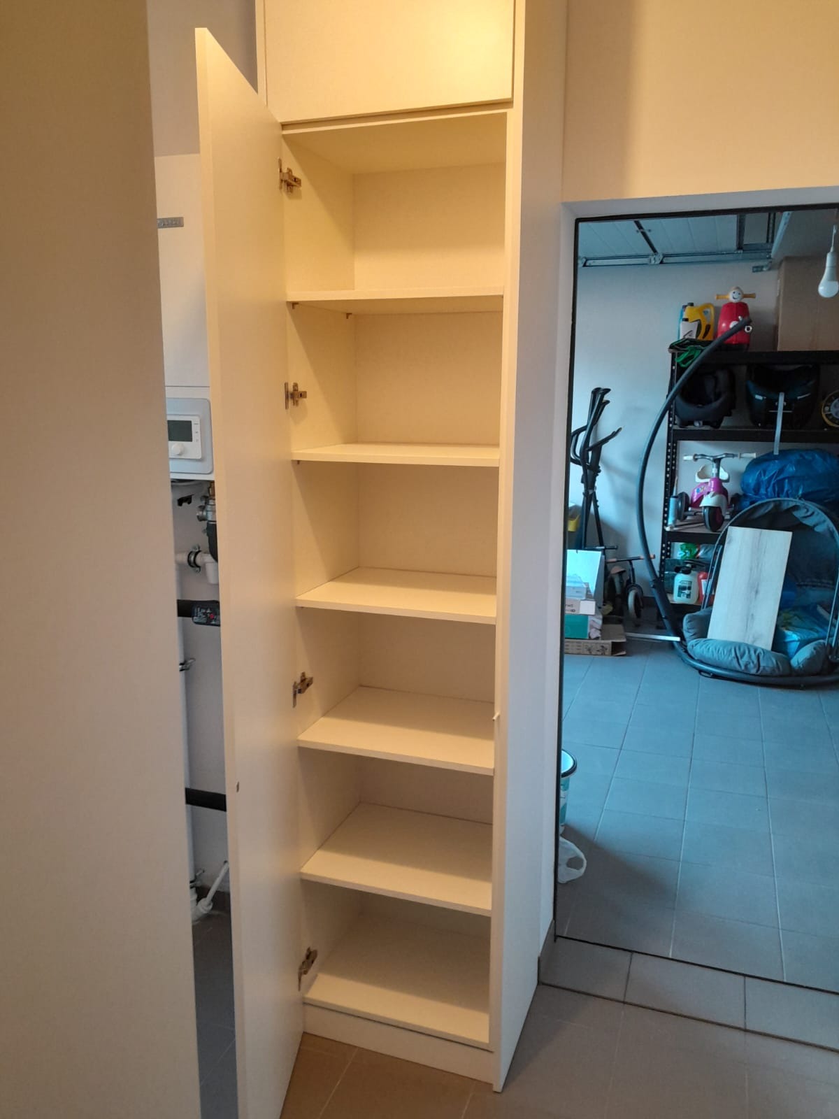 Wysoka, biała szafa z półkami, otwarte drzwi ukazują puste wnętrze, widok z boku na tle pomieszczenia gospodarczego.