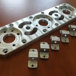 Aluminiowa płyta z precyzyjnie wyfrezowanymi otworami i zestaw małych, metalowych uchwytów na drewnianym blacie.