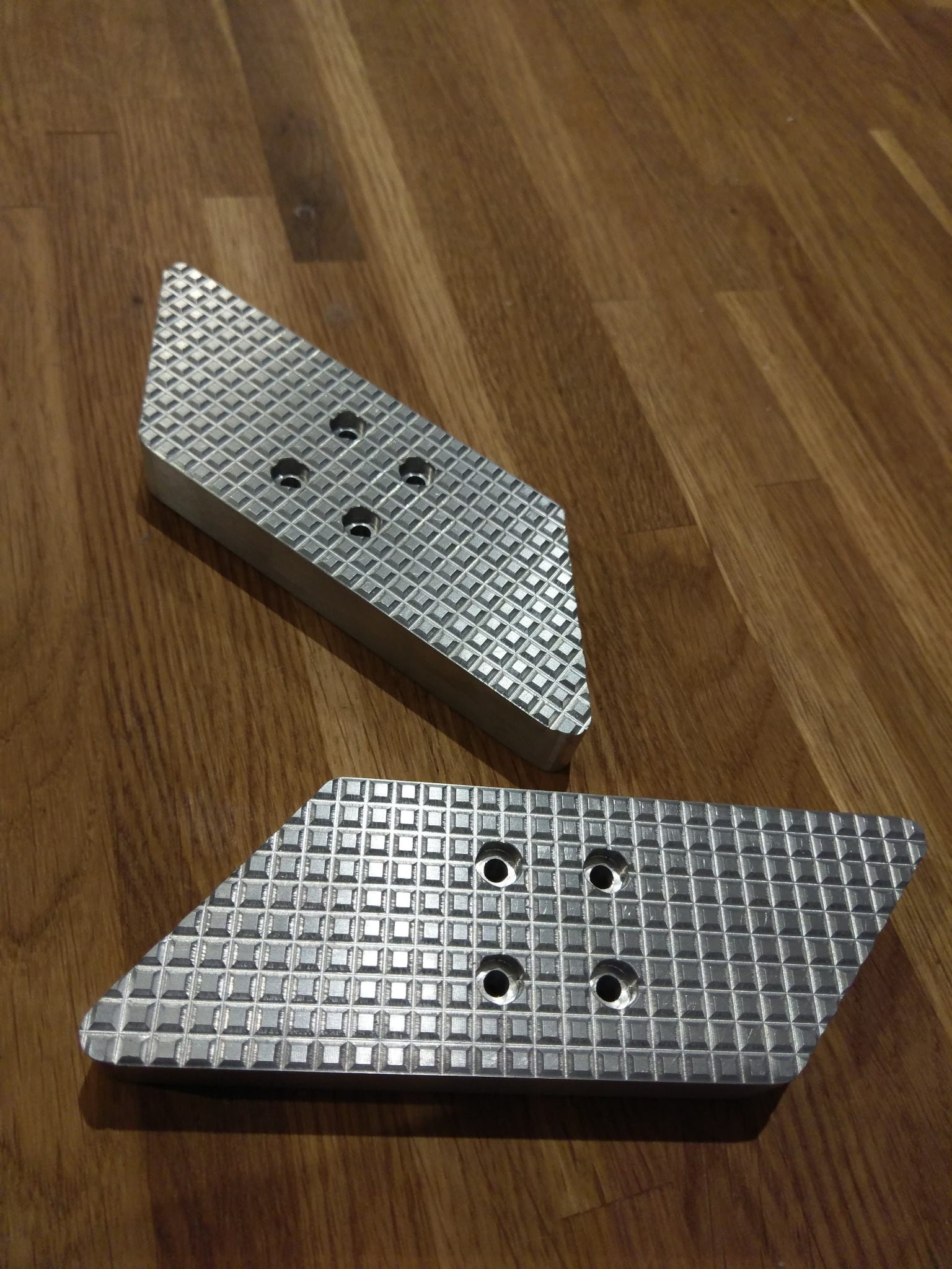 Dwa metalowe elementy o trapezoidalnym kształcie, z wytłoczoną kratką i otworami, leżące na drewnianej powierzchni.