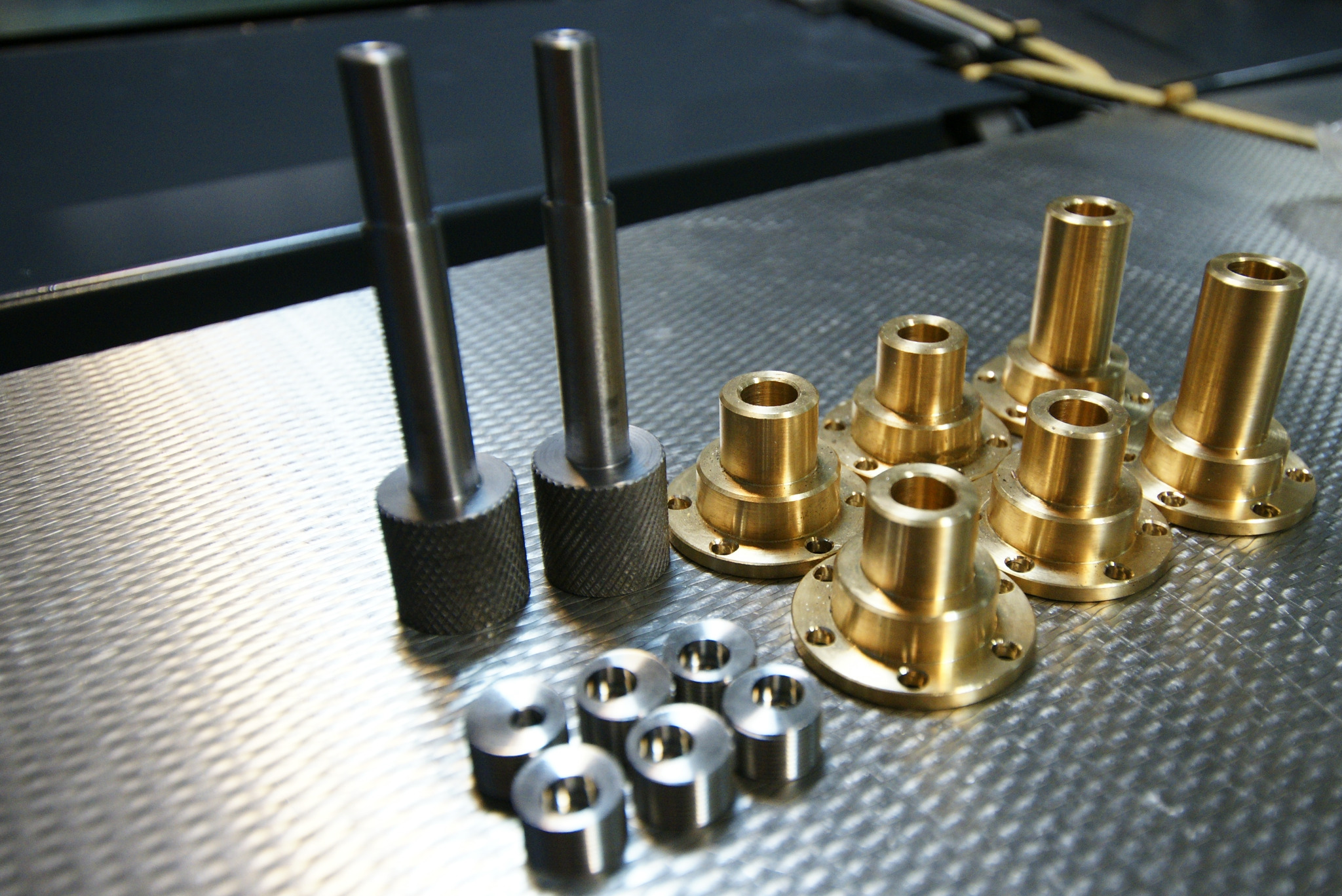 Precyzyjnie obrobione elementy metalowe: stalowe trzpienie z radełkowaną podstawą, mosiężne tuleje kołnierzowe i pierścienie, ułożone na metalowej powierzchni z widocznymi śladami obróbki.