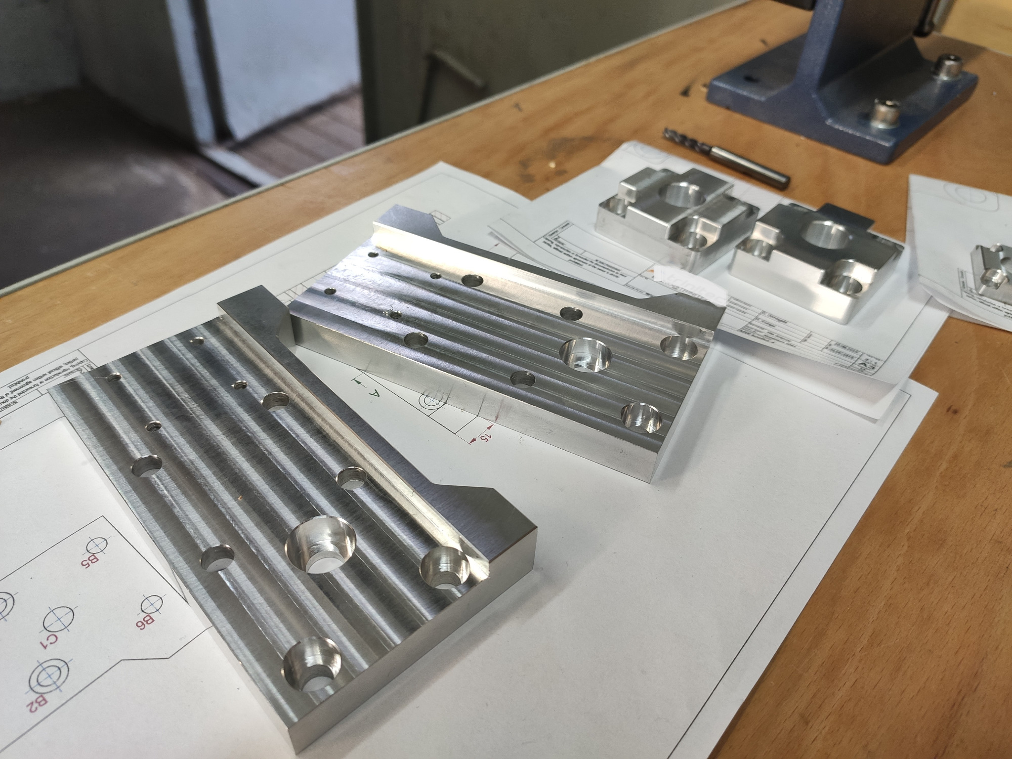 Metalowe elementy z otworami leżące na rysunku technicznym, widoczny frez i prasa śrubowa w tle.