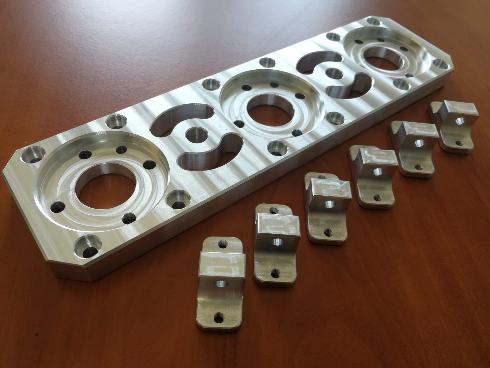 Aluminiowa płyta z precyzyjnie wyfrezowanymi otworami i zestaw małych, metalowych uchwytów na drewnianym blacie.