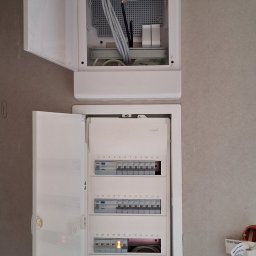 Usługi Elektryczne Emil Gajda - Dwa białe panele elektryczne zamontowane na szarej ścianie z otwartymi drzwiczkami, górny panel z kablem związanym czarną taśmą, dolny panel z bezpiecznikami, z których jeden jest włączony.