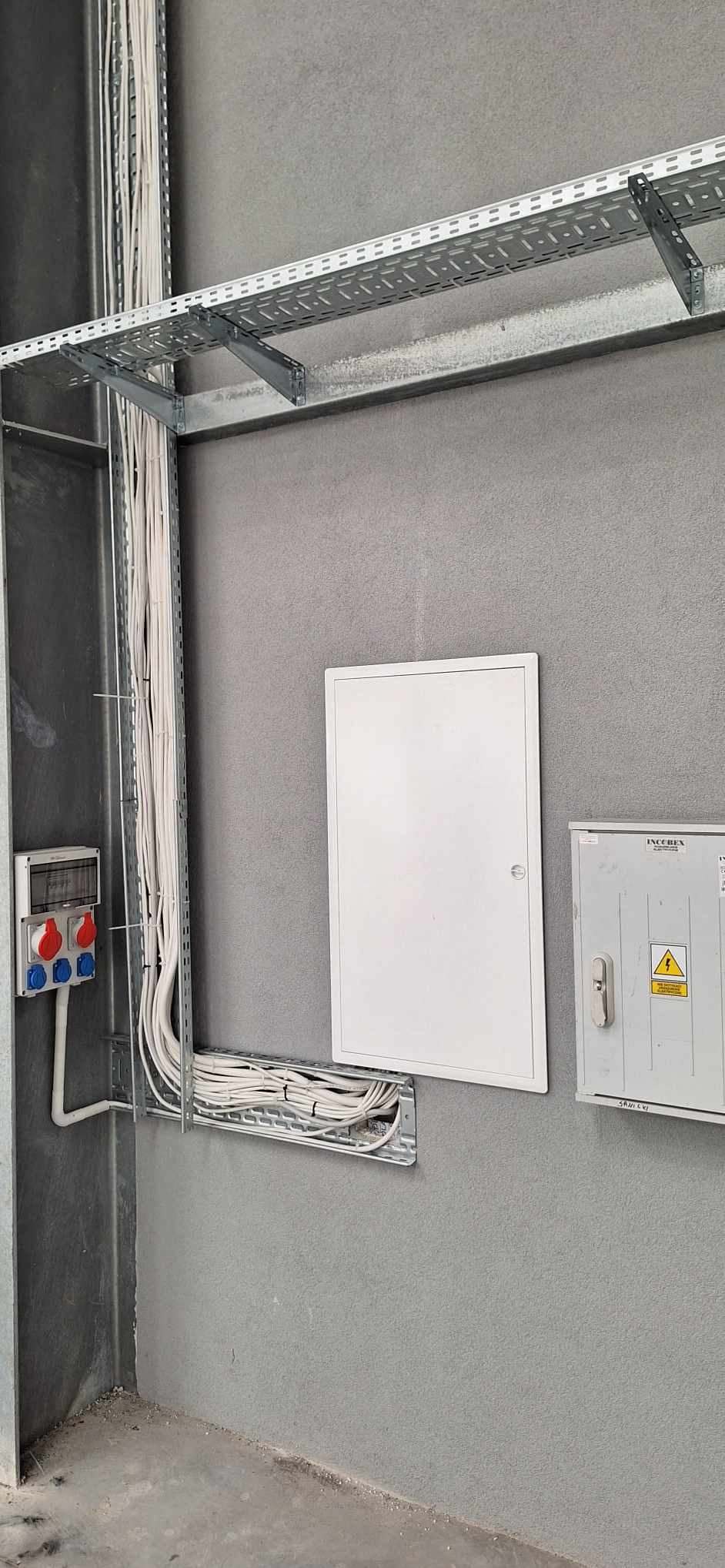 Szara ściana z zamontowanymi korytkami kablowymi, przewodami elektrycznymi, tablicą rozdzielczą z gniazdami siłowymi i skrzynką elektryczną z ostrzeżeniem o napięciu.
