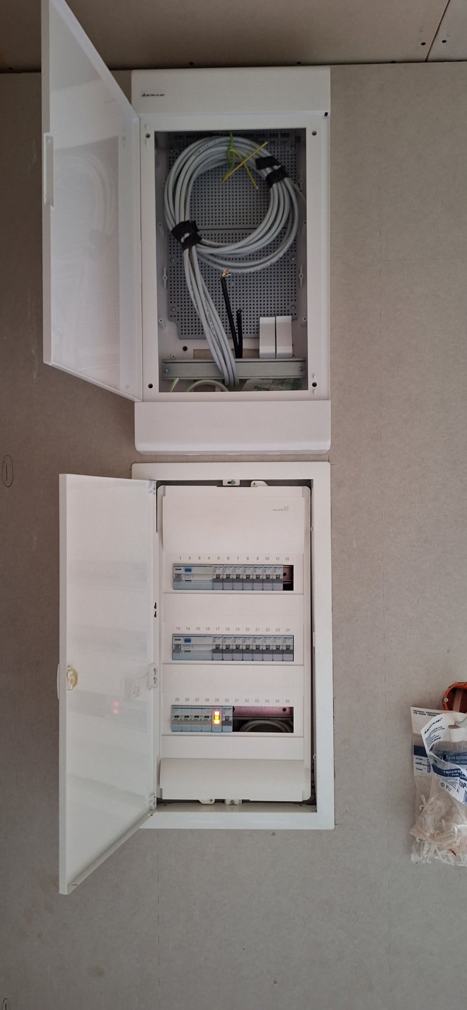 Dwa białe panele elektryczne zamontowane na szarej ścianie z otwartymi drzwiczkami, górny panel z kablem związanym czarną taśmą, dolny panel z bezpiecznikami, z których jeden jest włączony.