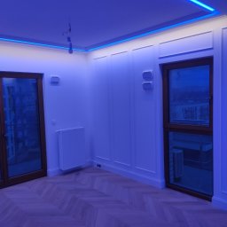 Likes Bud Andrii Fomich - Wykończone wnętrze pokoju z parkietem w jodełkę, białe ściany z dekoracyjnymi listwami i oknami. Sufit oświetlony niebieskimi ledami. Wisząca żarówka bez klosza.