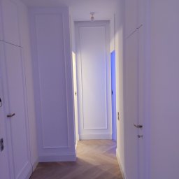 Likes Bud Andrii Fomich - Biały korytarz z parkietem ułożonym w jodełkę. Ściany z dekoracyjnymi listwami i wbudowanymi szafami. Oświetlenie punktowe na suficie.