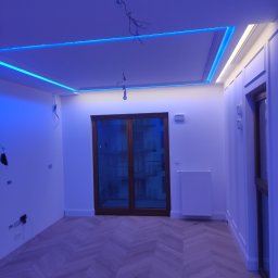 Likes Bud Andrii Fomich - Pokój z parkietem w jodełkę i oświetleniem LED na suficie. Widoczne okno balkonowe i białe ściany z dekoracyjnymi panelami. W tle grzejnik.