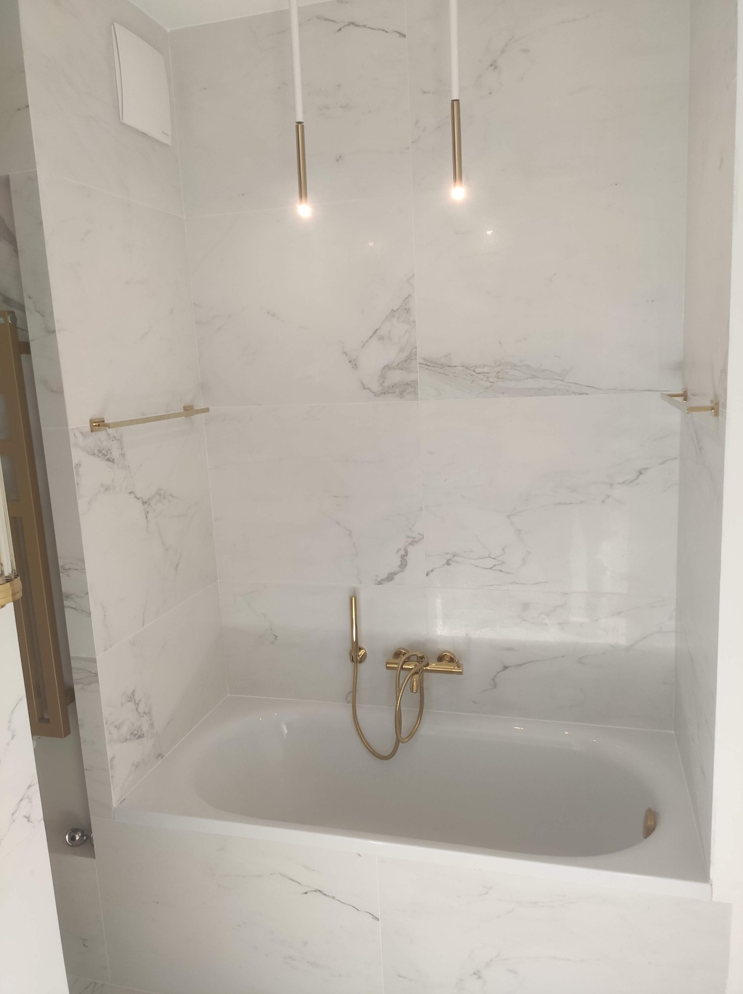 Biała łazienka z marmurowymi płytkami, złotą armaturą i minimalistycznymi lampami wiszącymi. Widok na wannę z baterią i relingami na ręczniki w kolorze złota.