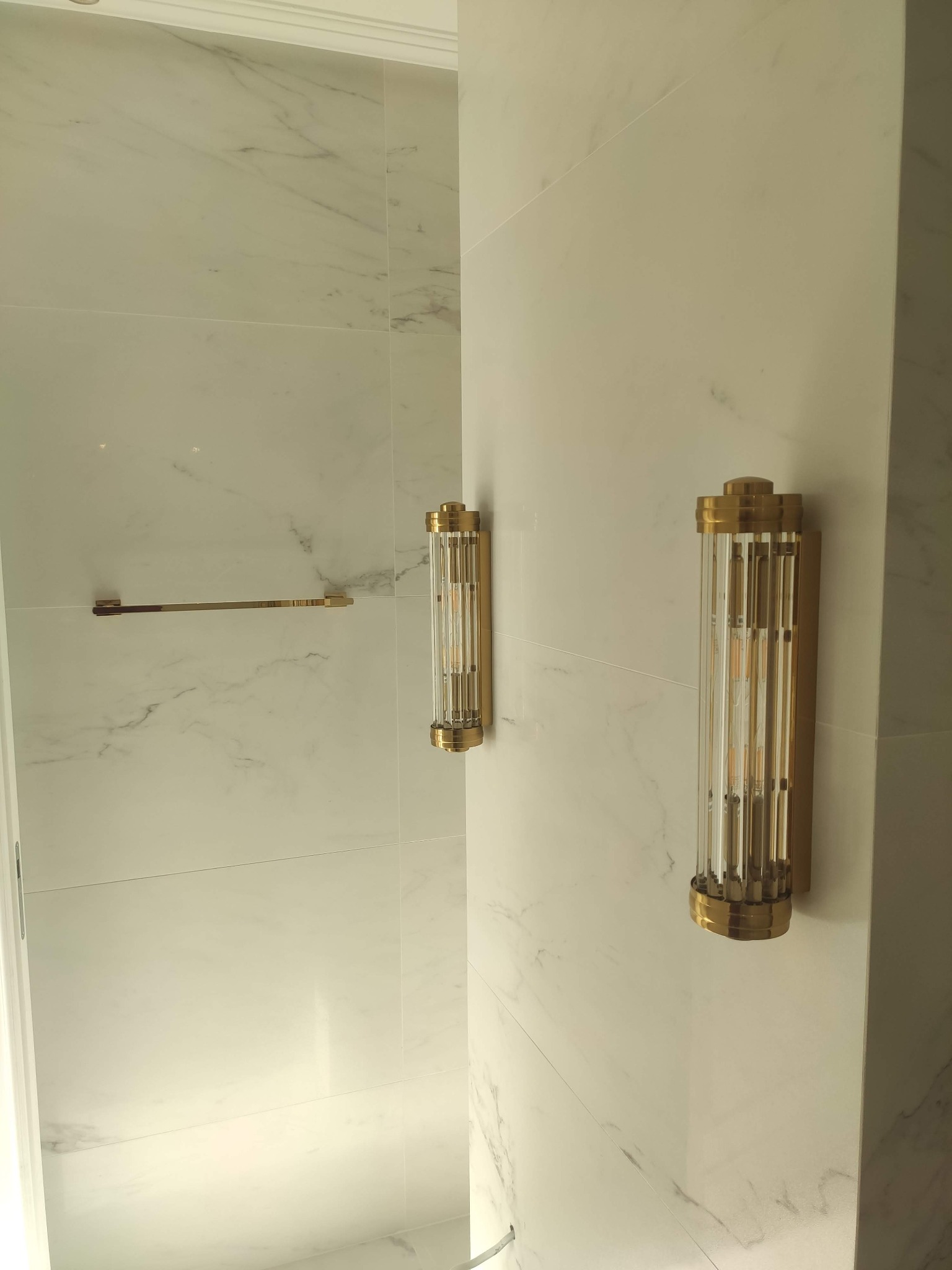 Luksusowa łazienka wykończona białym marmurem, z dwoma złotymi kinkietami i pasującym wieszakiem na ręczniki, tworząca elegancką i minimalistyczną przestrzeń.