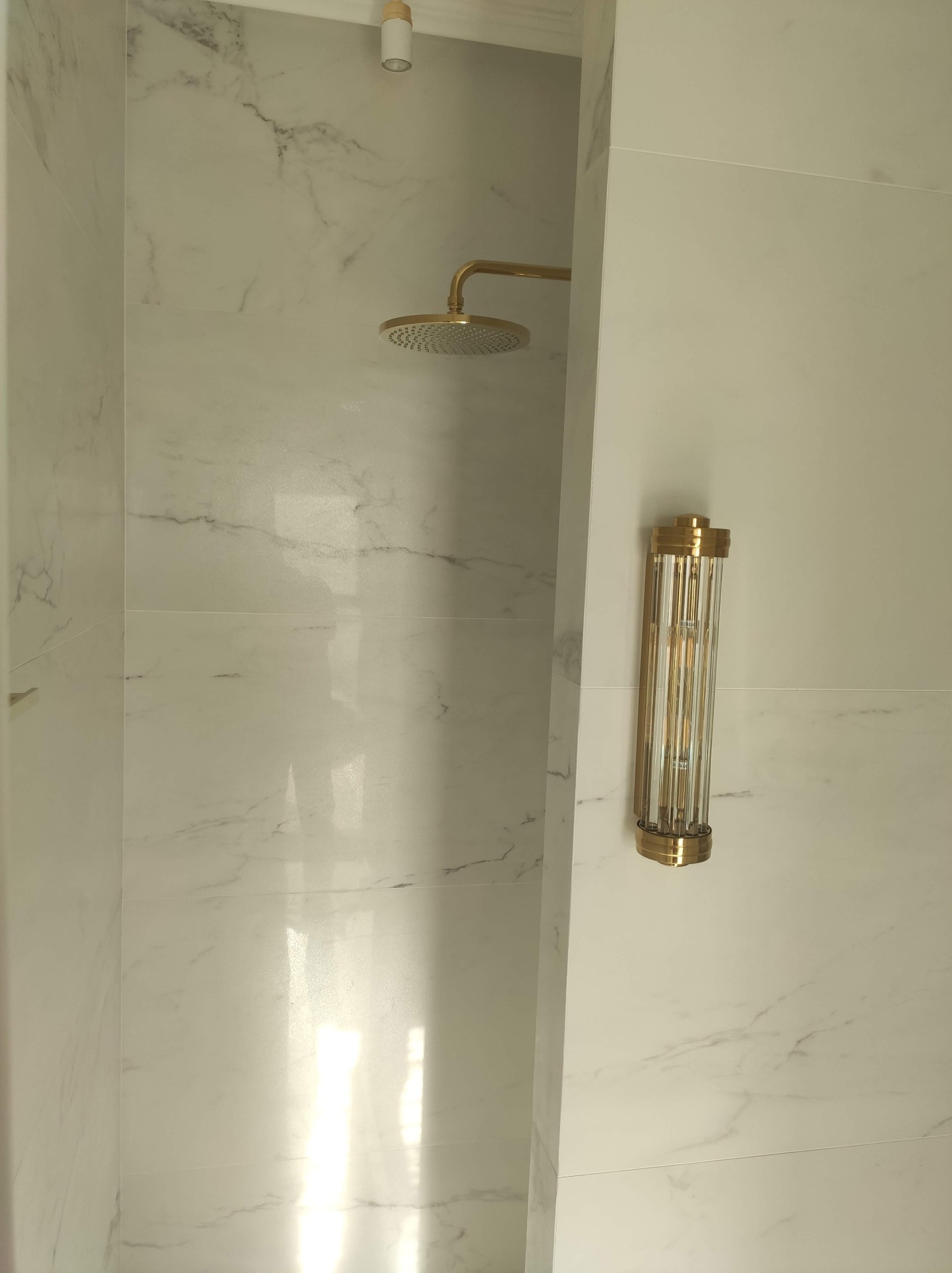 Luksusowa kabina prysznicowa z marmurowymi płytkami, złotą armaturą i ozdobnym kinkietem. Eleganckie wykończenie detali podkreśla wysoki standard.