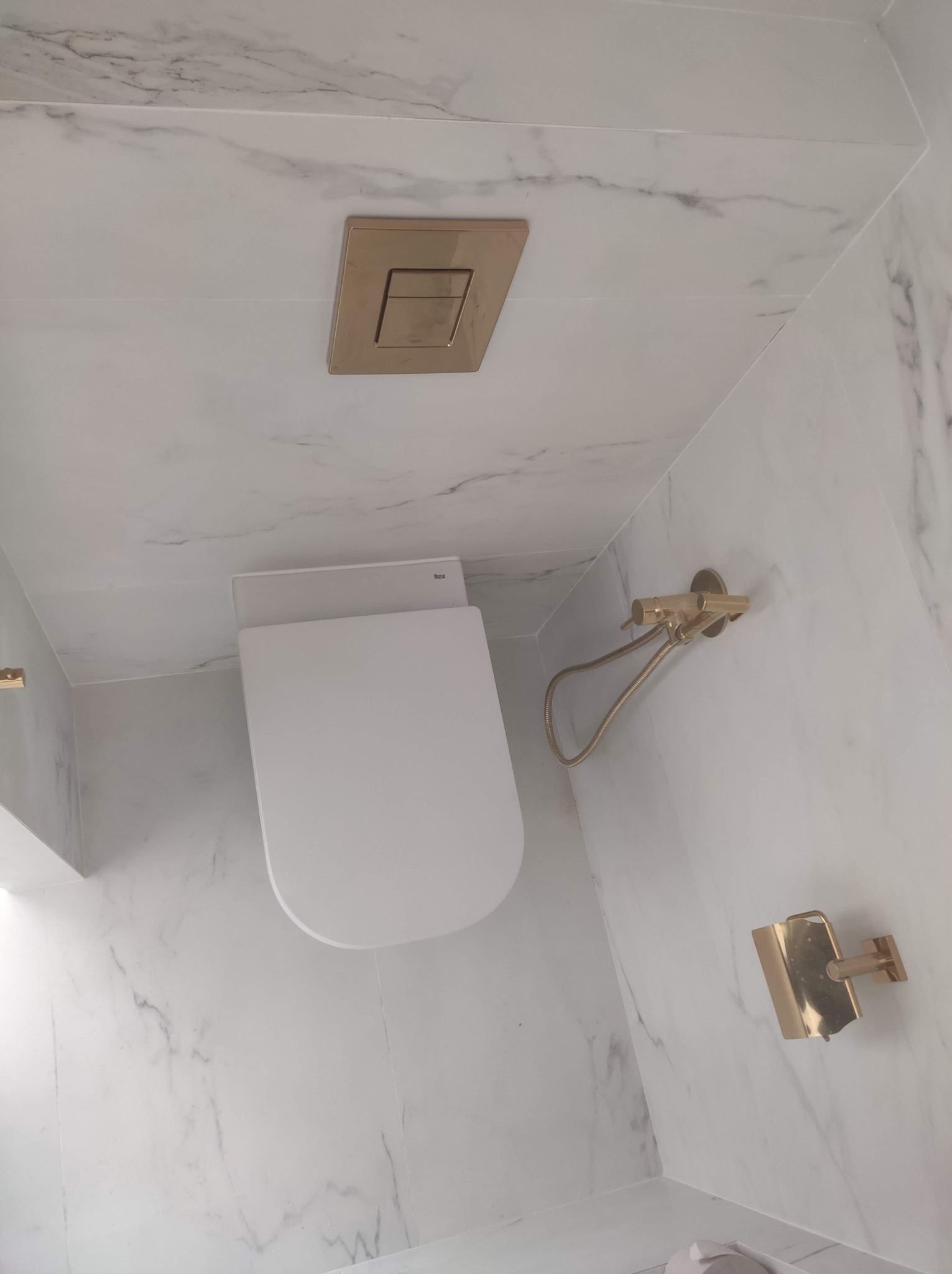 Nowoczesna łazienka z marmurowymi płytkami, wiszącą miską WC, złotą armaturą i przyciskiem spłukującym. Minimalistyczny design, eleganckie wykończenie.