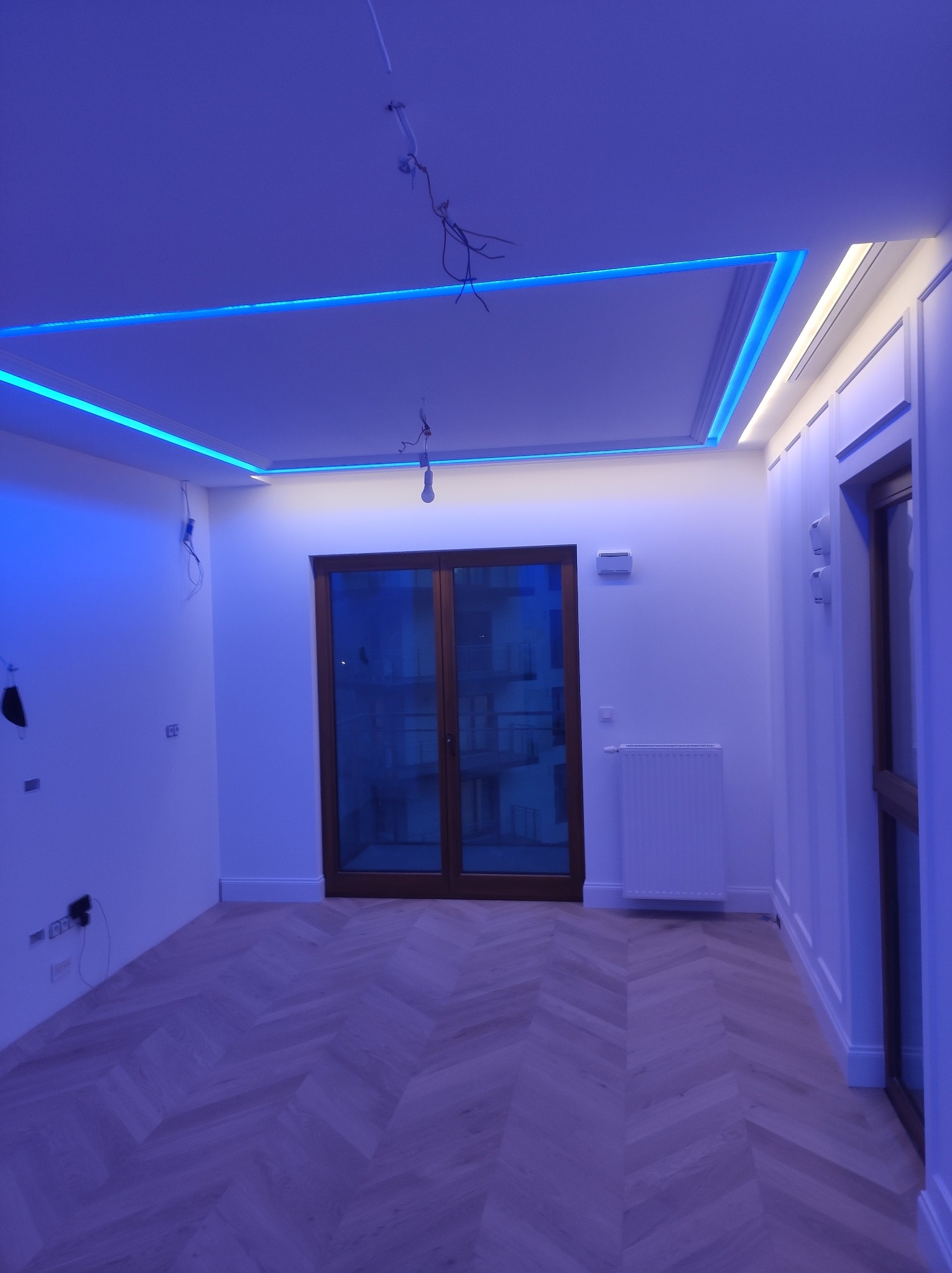 Pokój z parkietem w jodełkę i oświetleniem LED na suficie. Widoczne okno balkonowe i białe ściany z dekoracyjnymi panelami. W tle grzejnik.