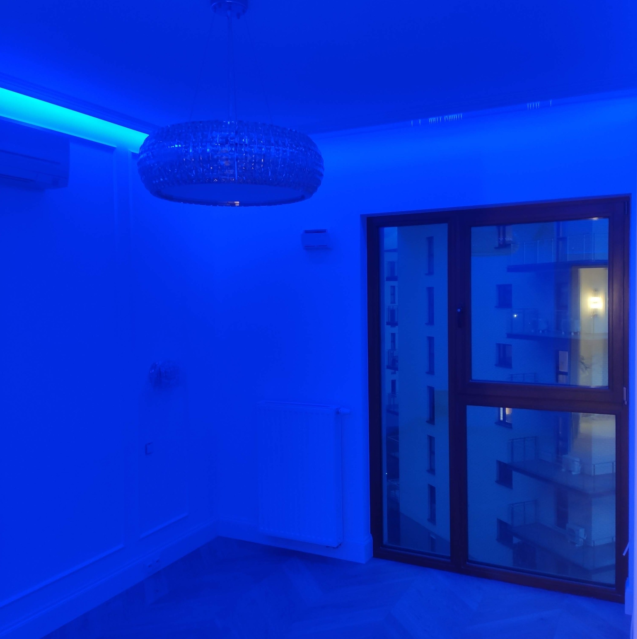 Pokój z oświetleniem LED, kryształowy żyrandol, okno z widokiem na budynek. Dominujący niebieski odcień nadaje nowoczesny charakter wnętrzu.