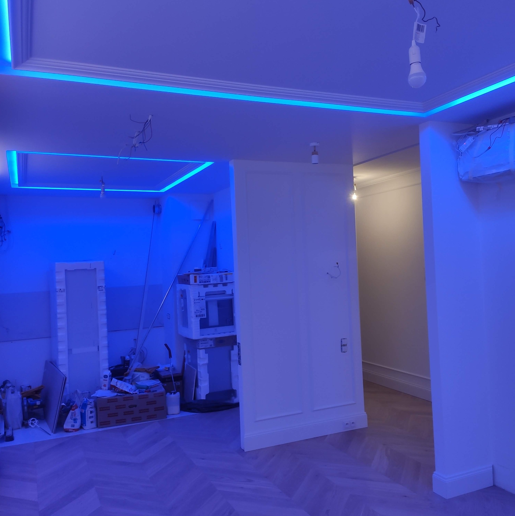 Niebieskie oświetlenie LED w suficie, dekoracyjne listwy, podłoga w jodełkę, widoczne prace wykończeniowe w tle, surowa żarówka, fragment klimatyzacji.