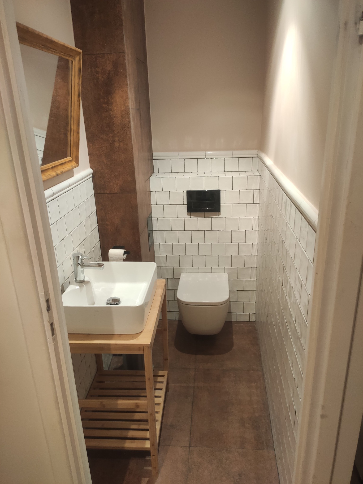 Kompaktowa łazienka z białą ceramiką i mozaiką. Umywalka na drewnianym stelażu, toaleta pod zabudowanym panelem. Lustro w złotej ramie dodaje elegancji. Całość w ciepłej tonacji.