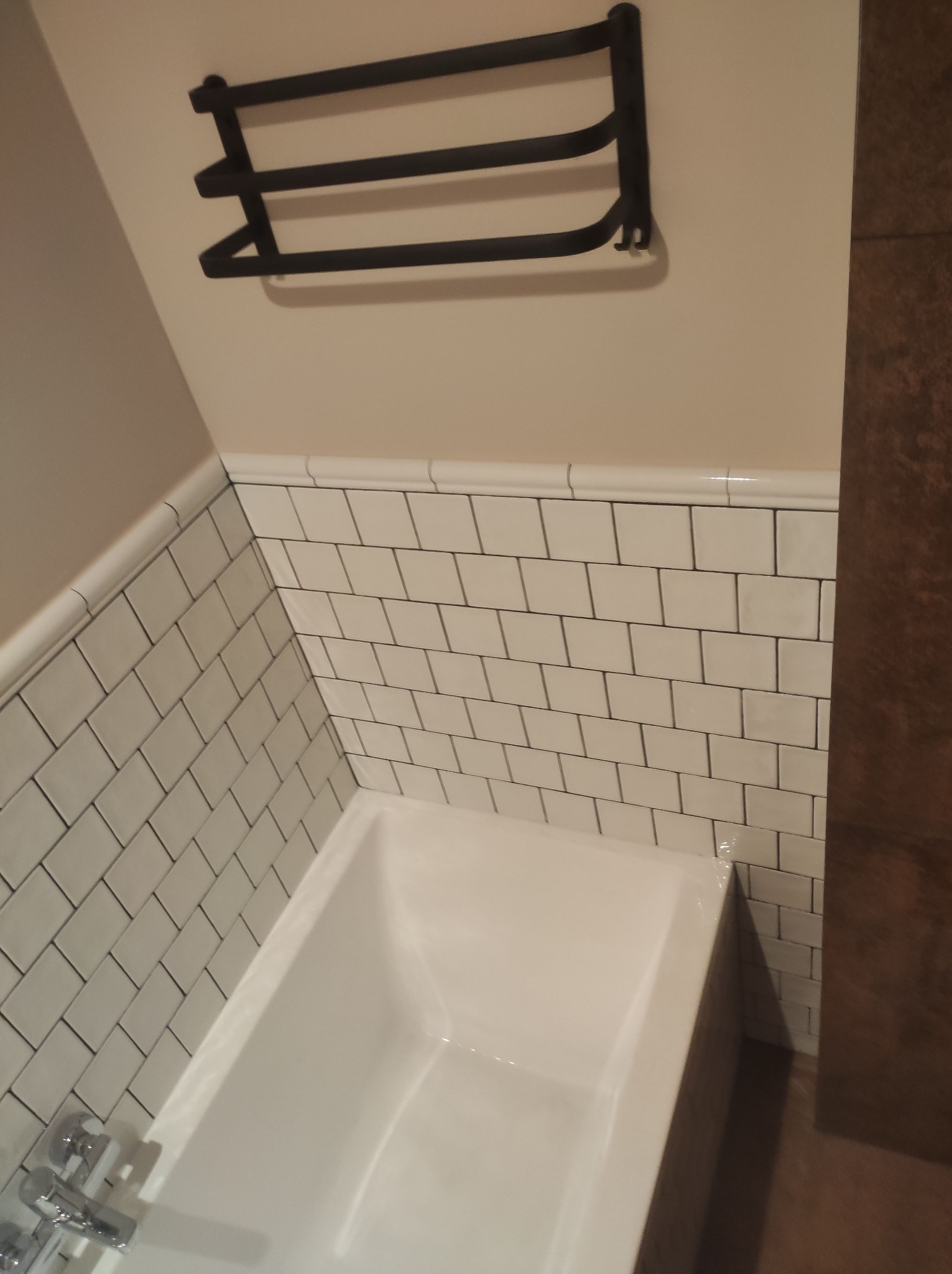 Biała wanna obłożona kafelkami typu metro, z czarnym, metalowym wieszakiem na ręczniki nad nią. Widoczny fragment armatury i drewniana obudowa wanny.