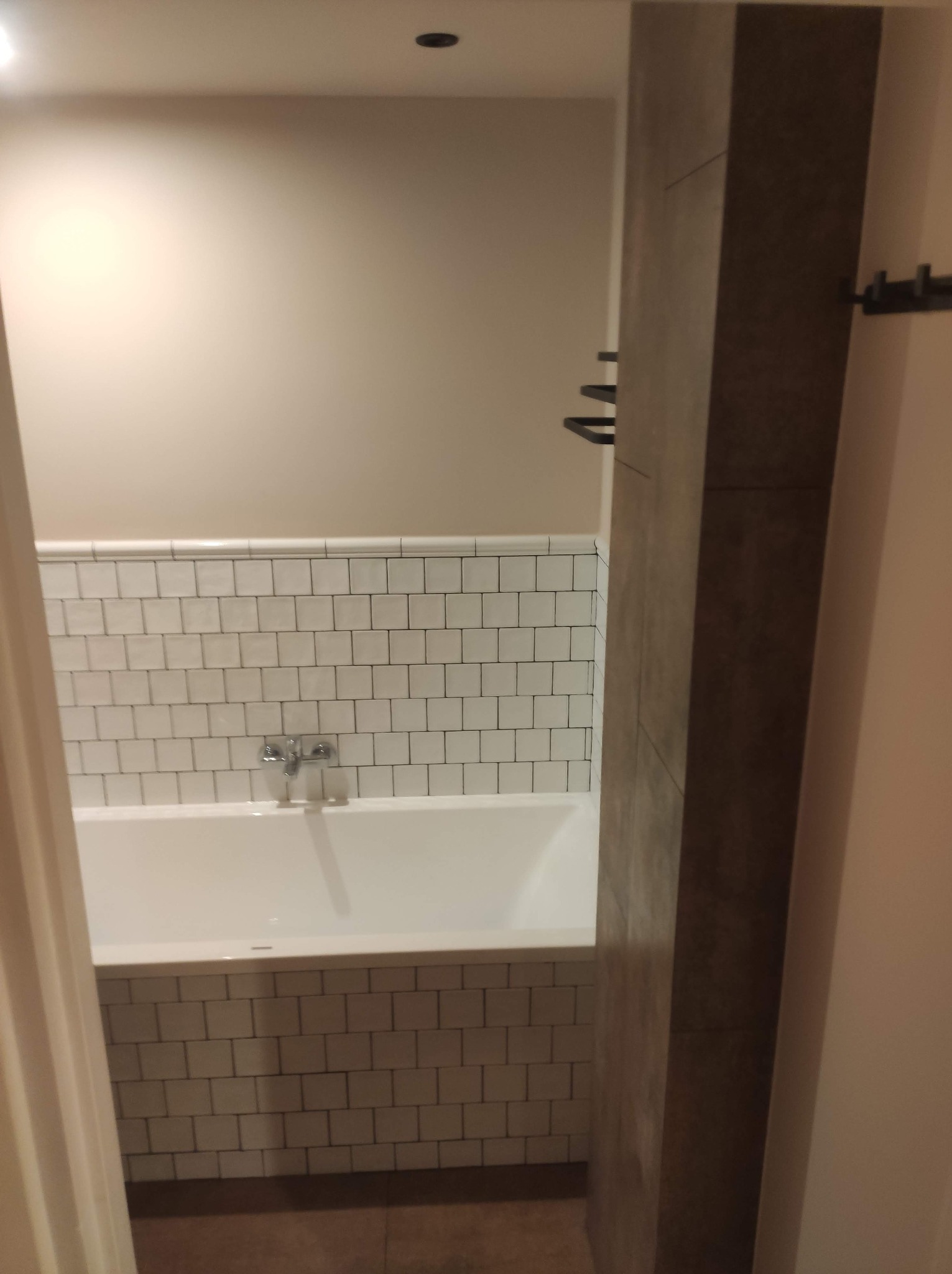 Biała wanna z baterią i obudową z płytek w małej łazience. Widok zza framugi drzwi.