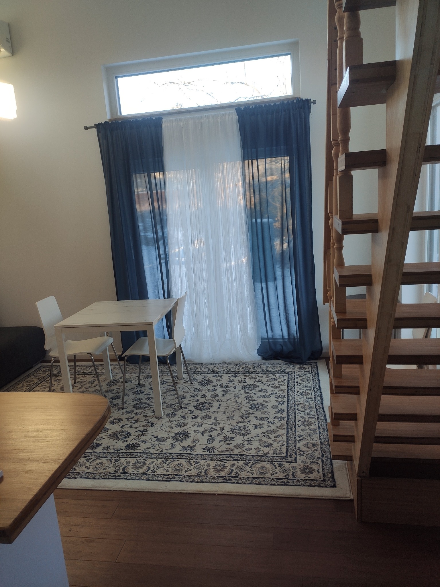 Jasne wnętrze z drewnianymi schodami, stołem i krzesłami na wzorzystym dywanie. Okno przesłonięte białymi i granatowymi zasłonami. Minimalistyczny design.