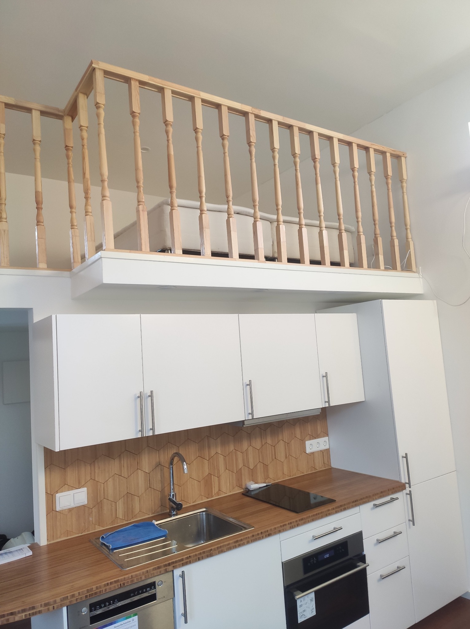 Nowoczesna kuchnia z antresolą sypialnianą: białe szafki, drewniany blat i heksagonalne płytki. Balustrada antresoli z jasnego drewna. Minimalistyczny design.