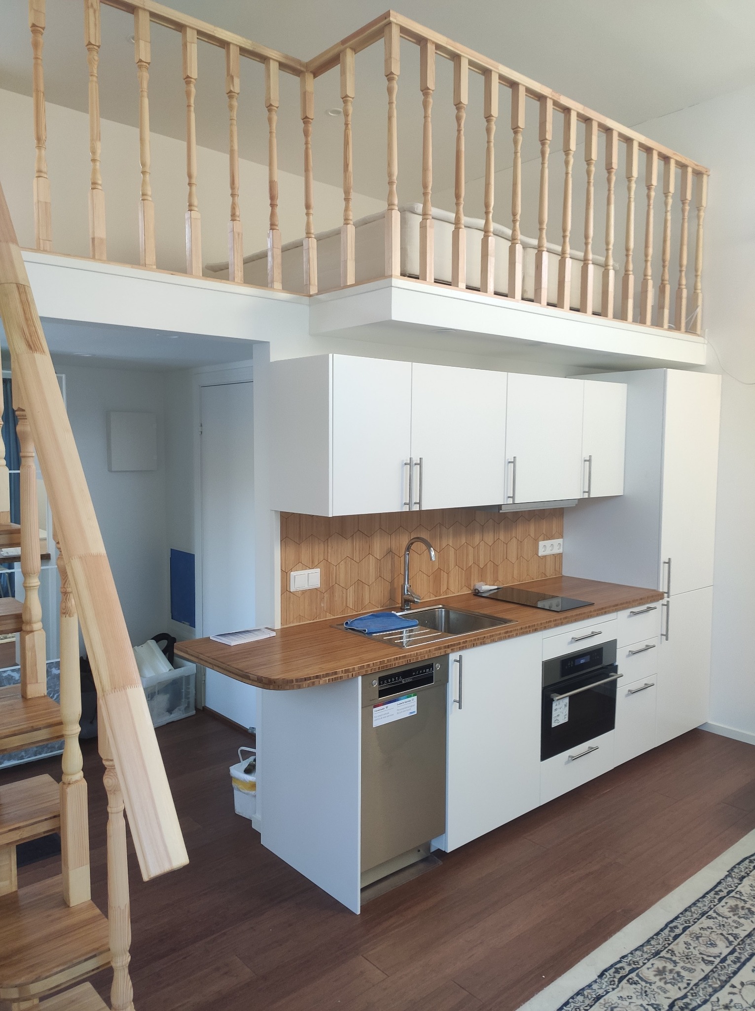 Nowoczesna kuchnia z drewnianym blatem i białymi szafkami. Nad kuchnią antresola z drewnianą balustradą. Widoczne schody, piekarnik i zlew.