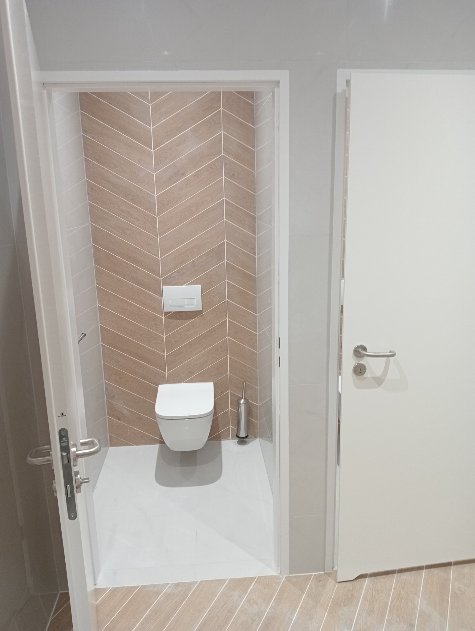 Nowoczesna łazienka z toaletą podwieszaną i wzorem jodełki na ścianie. Jasne płytki podłogowe i białe drzwi tworzą spójny, minimalistyczny design.