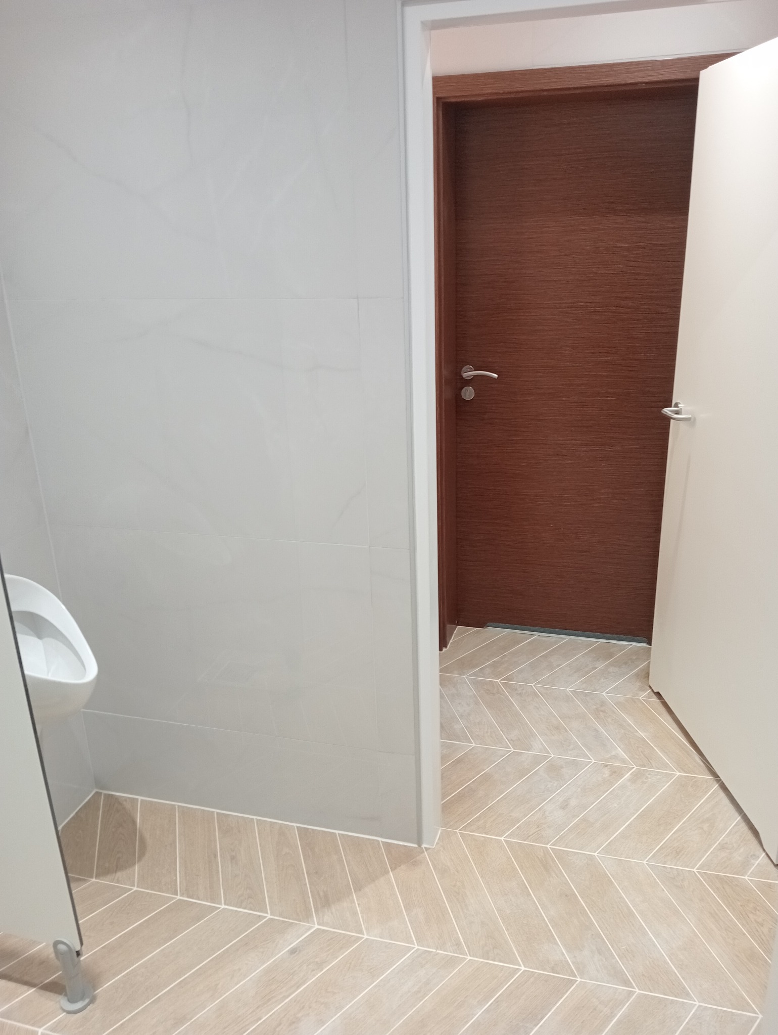 Nowoczesna łazienka publiczna z jasnymi płytkami ściennymi i podłogą w jodełkę. Widoczny pisuar i drewniane drzwi z metalową klamką. Minimalistyczny design.