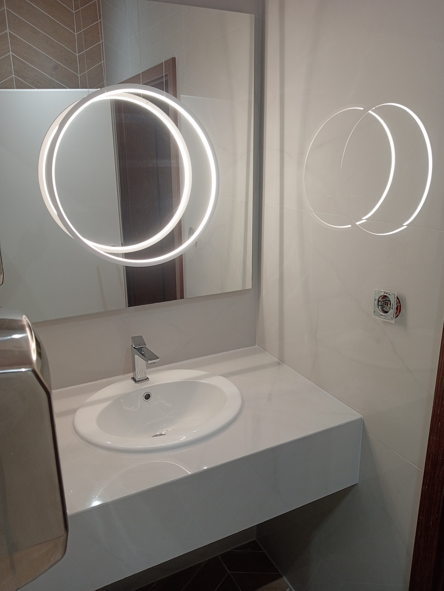 Nowoczesna łazienka z umywalką nablatową, okrągłym lustrem z podświetleniem LED i marmurowymi płytkami. Widoczny fragment instalacji elektrycznej.