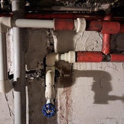 SZYMON JACIŃSKI - Instalacja hydrauliczna z rurami PCV i metalowymi, zawór kulowy z niebieskim pokrętłem, widoczne ślady po naprawach na ścianie.