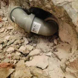 Szary kolanko kanalizacyjne HT osadzone w wykuciu w ścianie z gruzem, widoczna etykieta z kodem kreskowym.