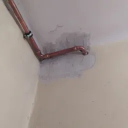 Miedziana rura instalacji gazowej lub wodnej, zamontowana w rogu pomieszczenia, z widocznymi śladami zaprawy i niedomalowaną ścianą.