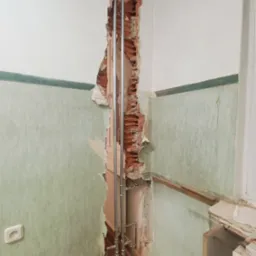 Odkryte rury instalacji grzewczej w bruździe ściany z fragmentami starej, zielonej tapety i odsłoniętą cegłą, widoczny grzejnik panelowy.