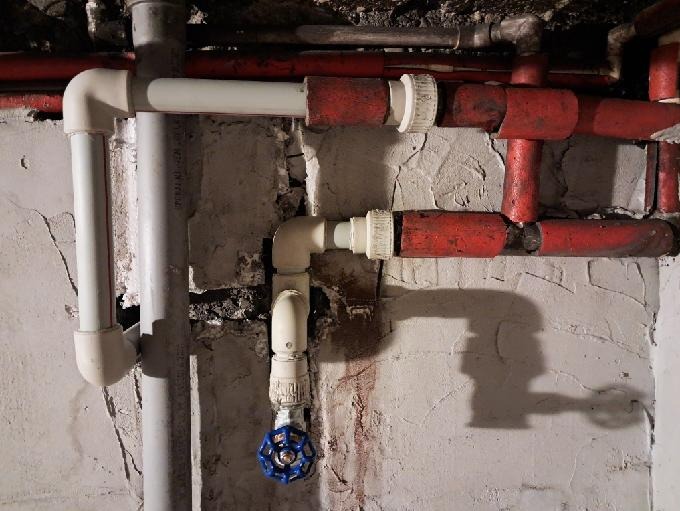 Instalacja hydrauliczna z rurami PCV i metalowymi, zawór kulowy z niebieskim pokrętłem, widoczne ślady po naprawach na ścianie.