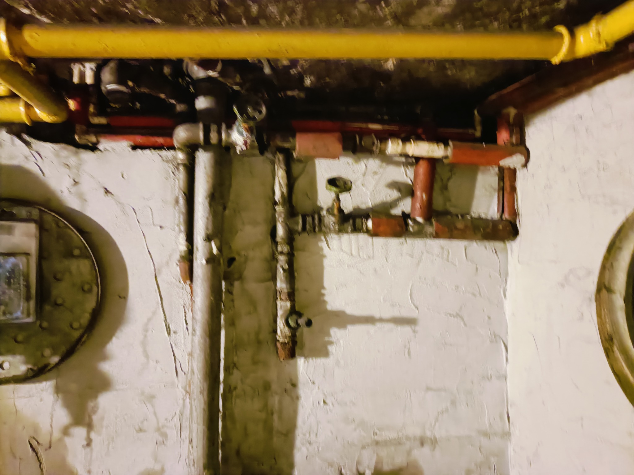 Fragment piwnicy z rurami gazowymi i wodnymi, żółte rury gazowe biegnące pod sufitem, czerwone i szare rury wodne na ścianie z grubym tynkiem, widoczne zawory i łączenia, oświetlenie górne.