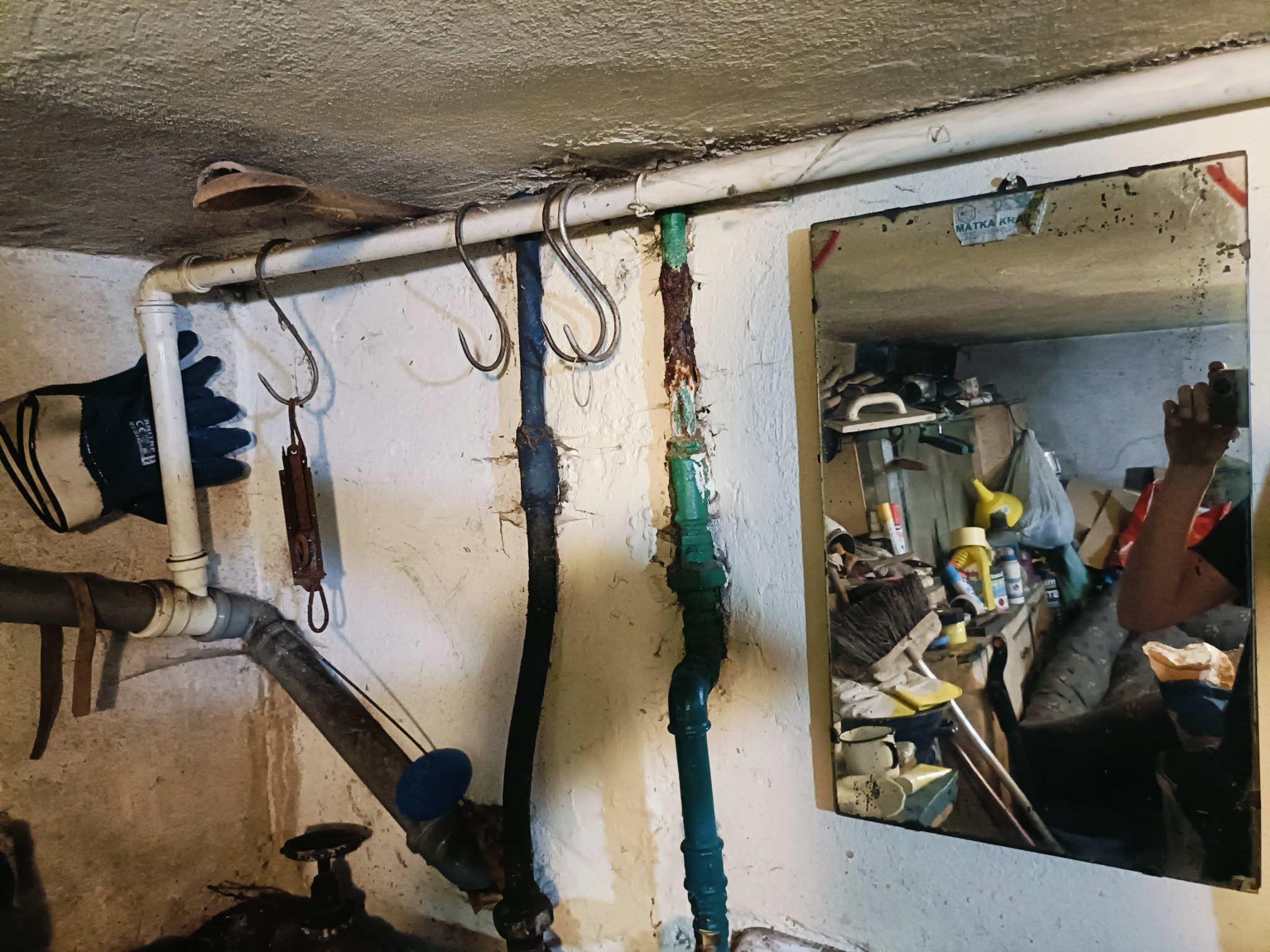 Stare, zardzewiałe rury w piwnicy, widoczne haki na rurze, rękawica robocza i brudne lustro odbijające zagracone pomieszczenie.