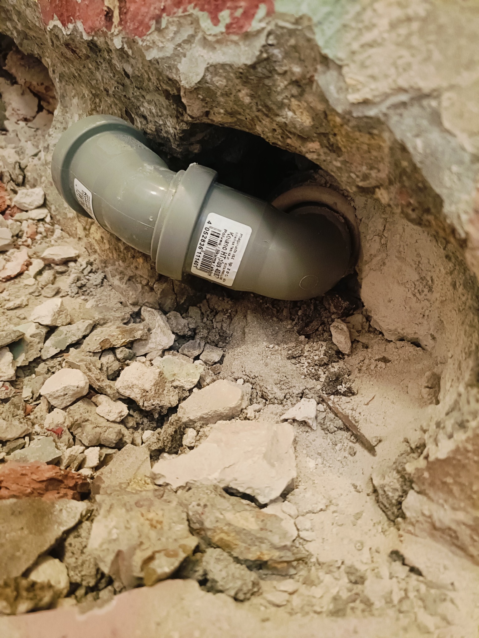 Szary kolanko kanalizacyjne HT osadzone w wykuciu w ścianie z gruzem, widoczna etykieta z kodem kreskowym.