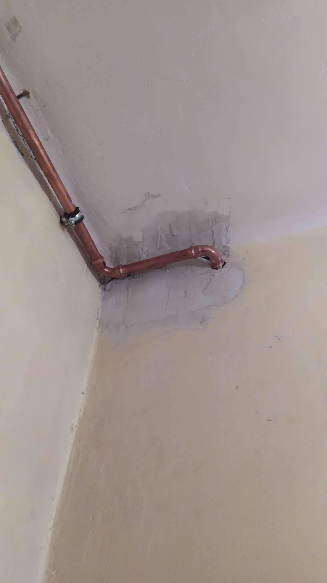 Miedziana rura instalacji gazowej lub wodnej, zamontowana w rogu pomieszczenia, z widocznymi śladami zaprawy i niedomalowaną ścianą.
