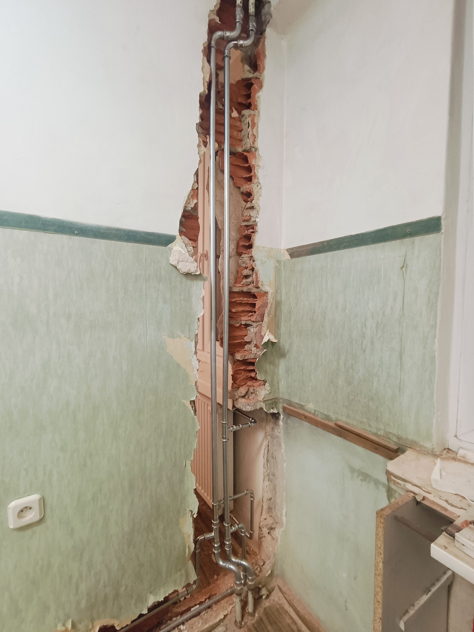 Odkryte rury instalacji grzewczej w bruździe ściany z fragmentami starej, zielonej tapety i odsłoniętą cegłą, widoczny grzejnik panelowy.