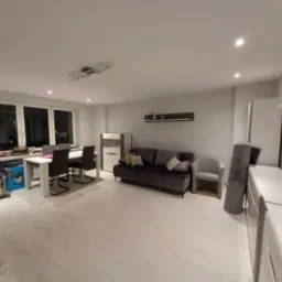 Jasne przestronne wnętrze z białymi ścianami i podłogą, szarą sofą i stołem jadalnym, widoczny sprzęt budowlany w rogu pokoju.