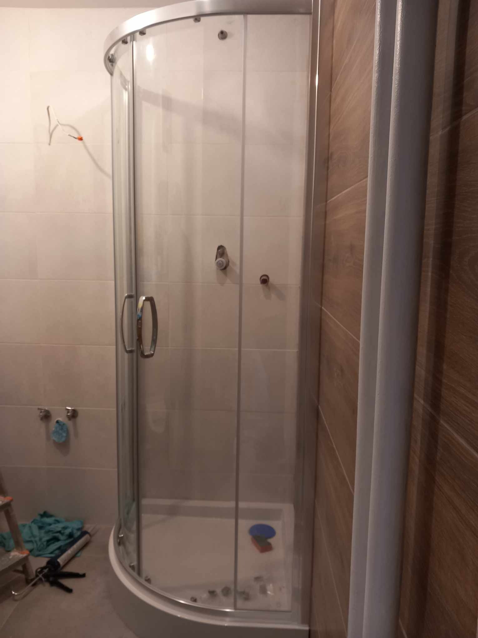 Narożna kabina prysznicowa z transparentnymi drzwiami i białym brodzikiem, zamontowana w łazience z beżowymi płytkami i drewnopodobną ścianą, widoczne narzędzia i drabina sugerują prace wykończeniowe.