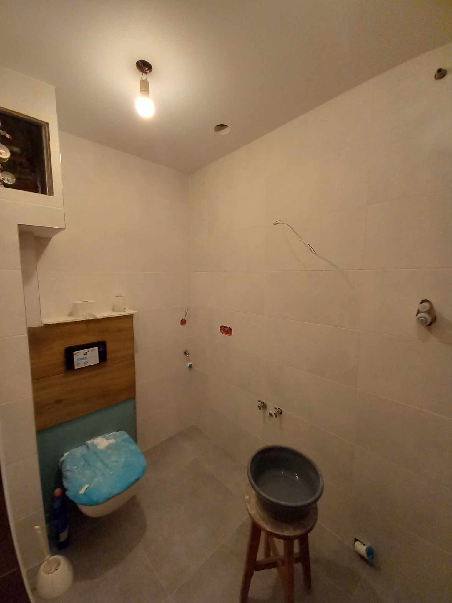 Wnętrze łazienki w trakcie remontu: białe płytki na ścianach, szary gres na podłodze, wisząca miska WC zabezpieczona folią, drewniana zabudowa stelaża, umywalka na drewnianym stołku, widoczne...