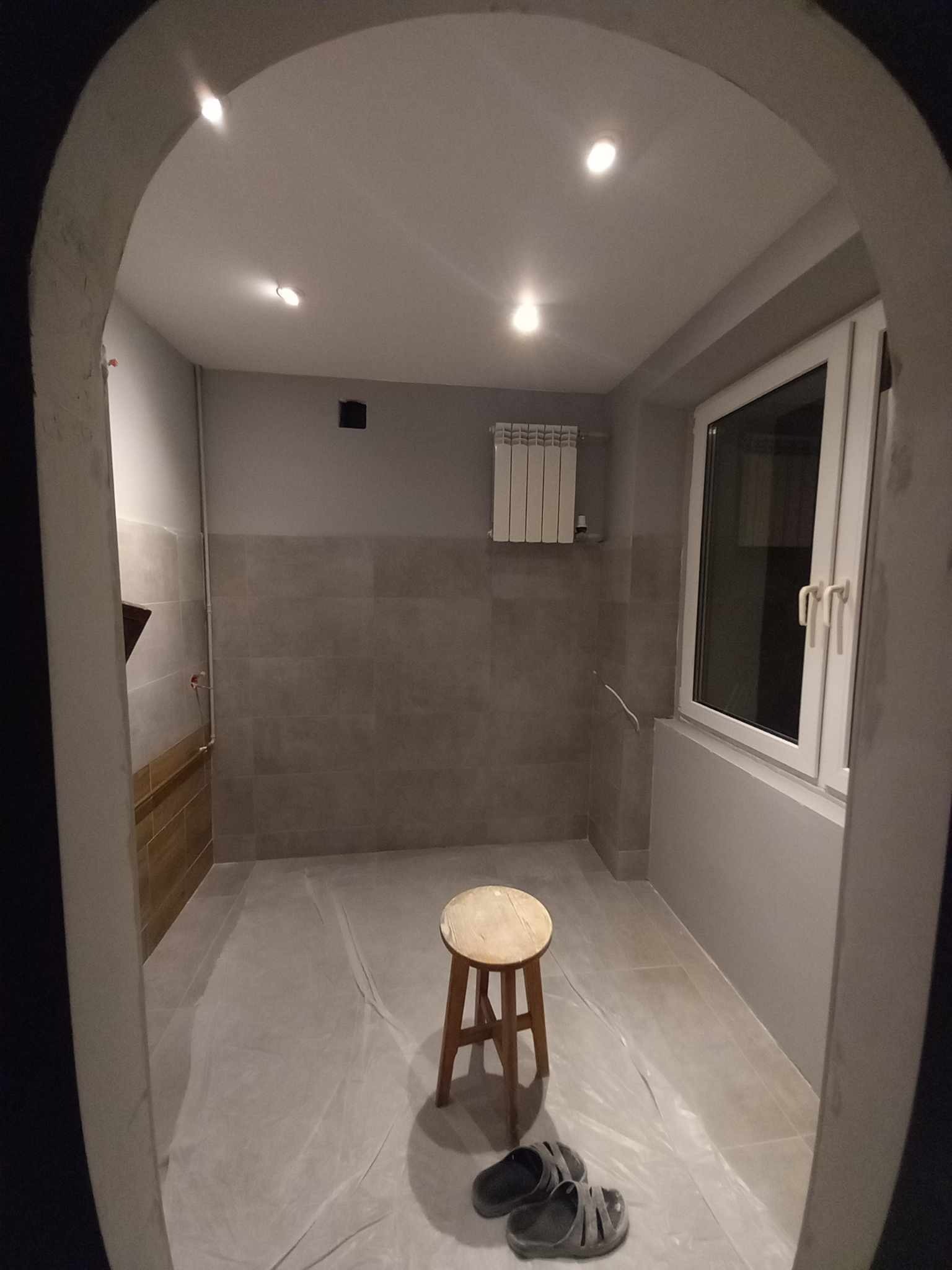Wnętrze łazienki w trakcie remontu, z zabezpieczoną folią podłogą, drewnianym stołkiem, grzejnikiem i widocznymi płytkami na ścianach. Oświetlenie sufitowe punktowe.