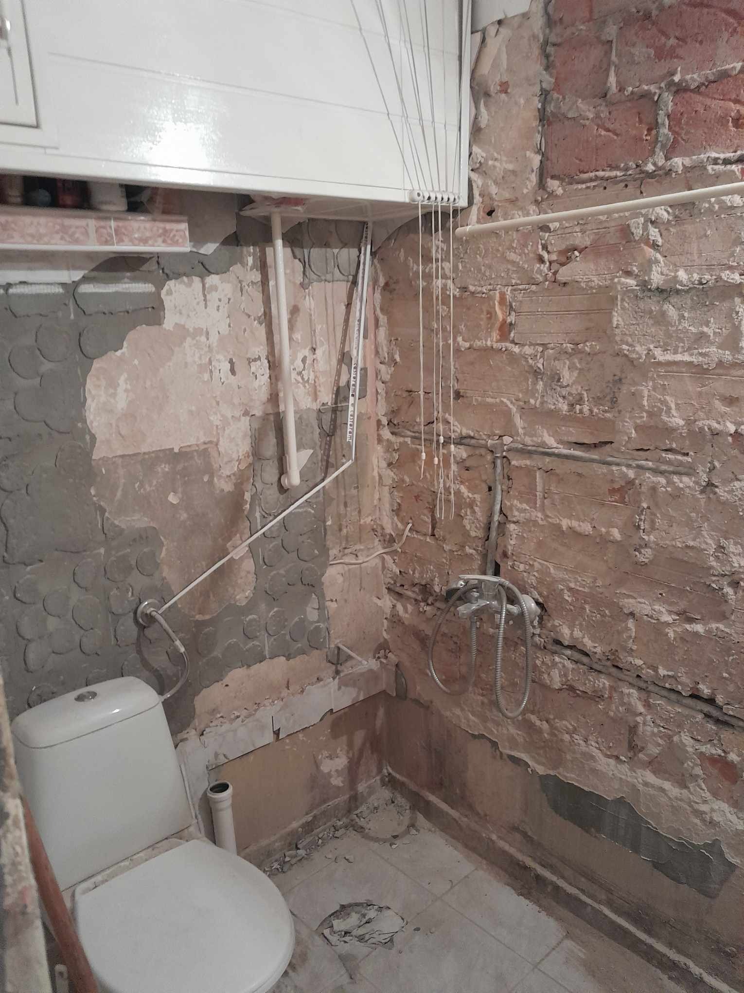 Łazienka w trakcie remontu: odsłonięta cegła, częściowo położona gładź, widoczne rury, zamontowana toaleta i suszarka podsufitowa.