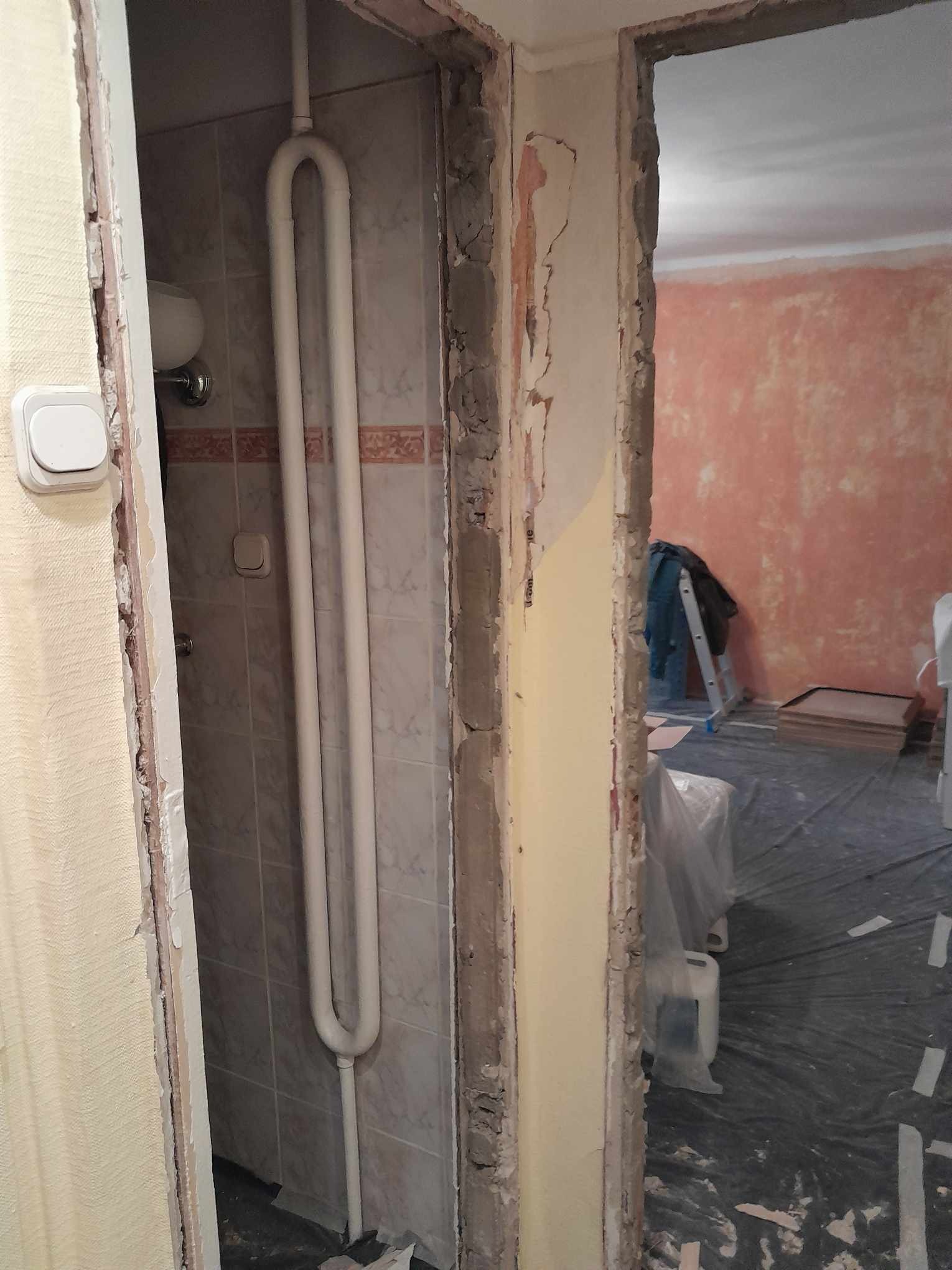 Łazienka w trakcie demontażu: widoczne płytki, grzejnik drabinkowy oraz ogołocone futryny drzwi, w tle zabezpieczona folią podłoga i drabina.