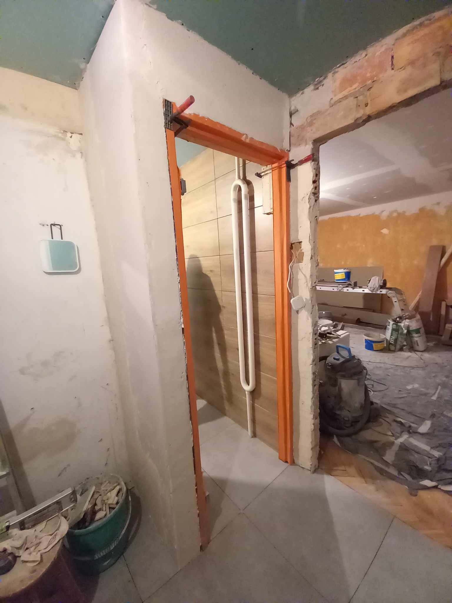 Wnętrze łazienki w trakcie remontu: widoczna rama drzwiowa pomalowana na pomarańczowo, grzejnik drabinkowy na ścianie wyłożonej płytkami oraz nieotynkowane ściany i podłoga w trakcie prac...