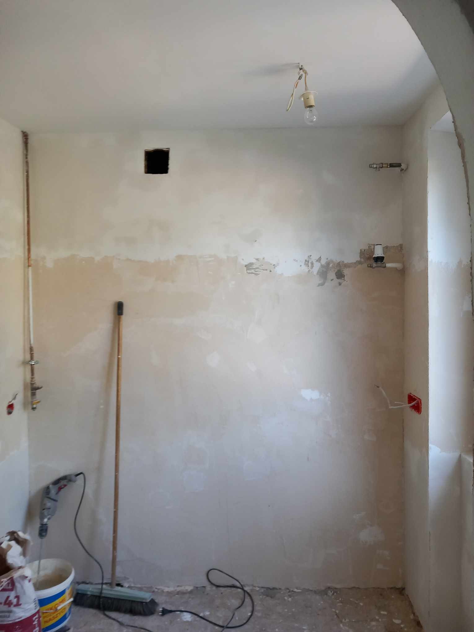 Pomieszczenie w trakcie prac wykończeniowych: ściany częściowo pokryte gładzią, widoczne instalacje wodne, elektryczne, wisząca żarówka na kablu, wiadro z zaprawą, wiertarka i miotła.