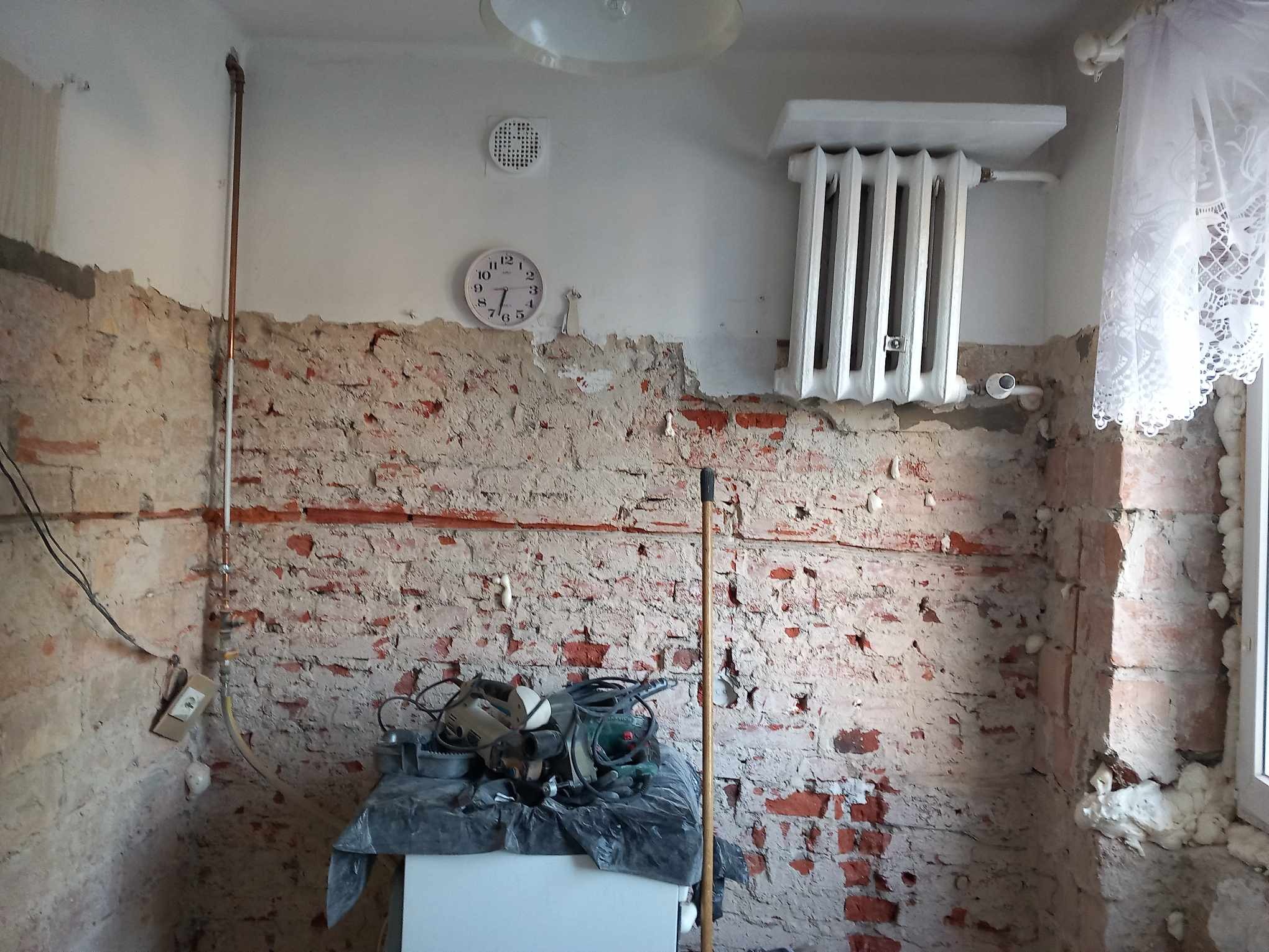 Częściowo otynkowana ściana z odsłoniętą czerwoną cegłą w trakcie remontu, widoczne narzędzia na stole przy ścianie, grzejnik na ścianie, zegar i lampa sufitowa.