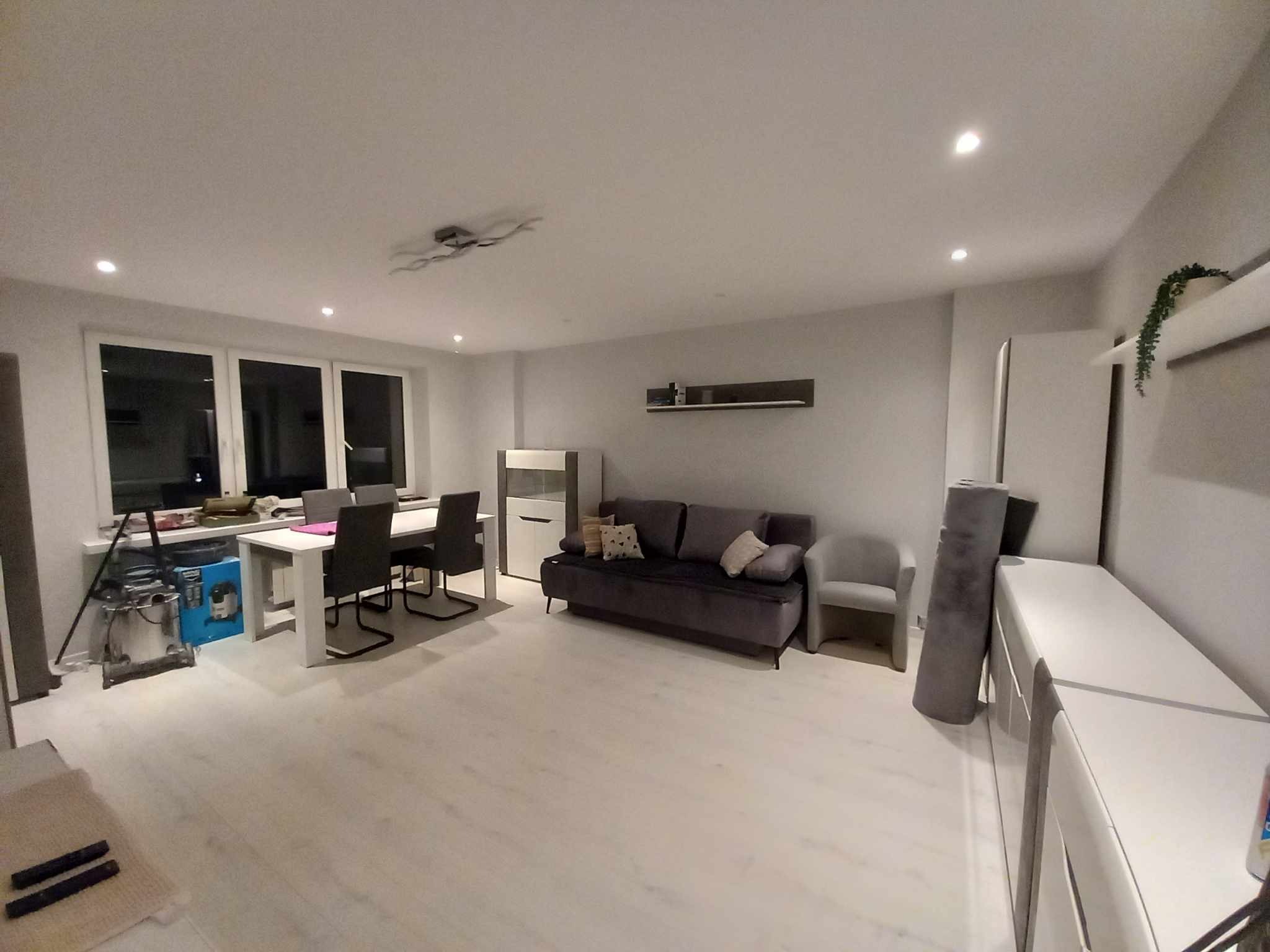 Jasne przestronne wnętrze z białymi ścianami i podłogą, szarą sofą i stołem jadalnym, widoczny sprzęt budowlany w rogu pokoju.