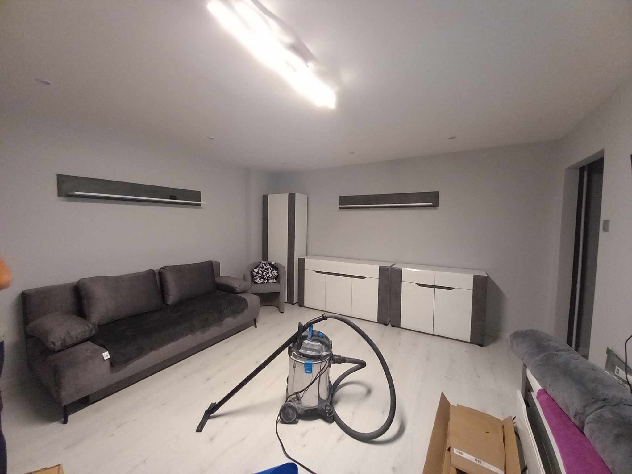 Wnętrze pokoju po remoncie z szarą sofą, biało-szarą komodą, grzejnikami panelowymi pod sufitem i odkurzaczem przemysłowym na podłodze.